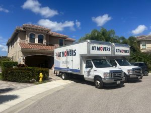 Moving company trucks parked in front of a residential two-story house in a suburban neighborhood.