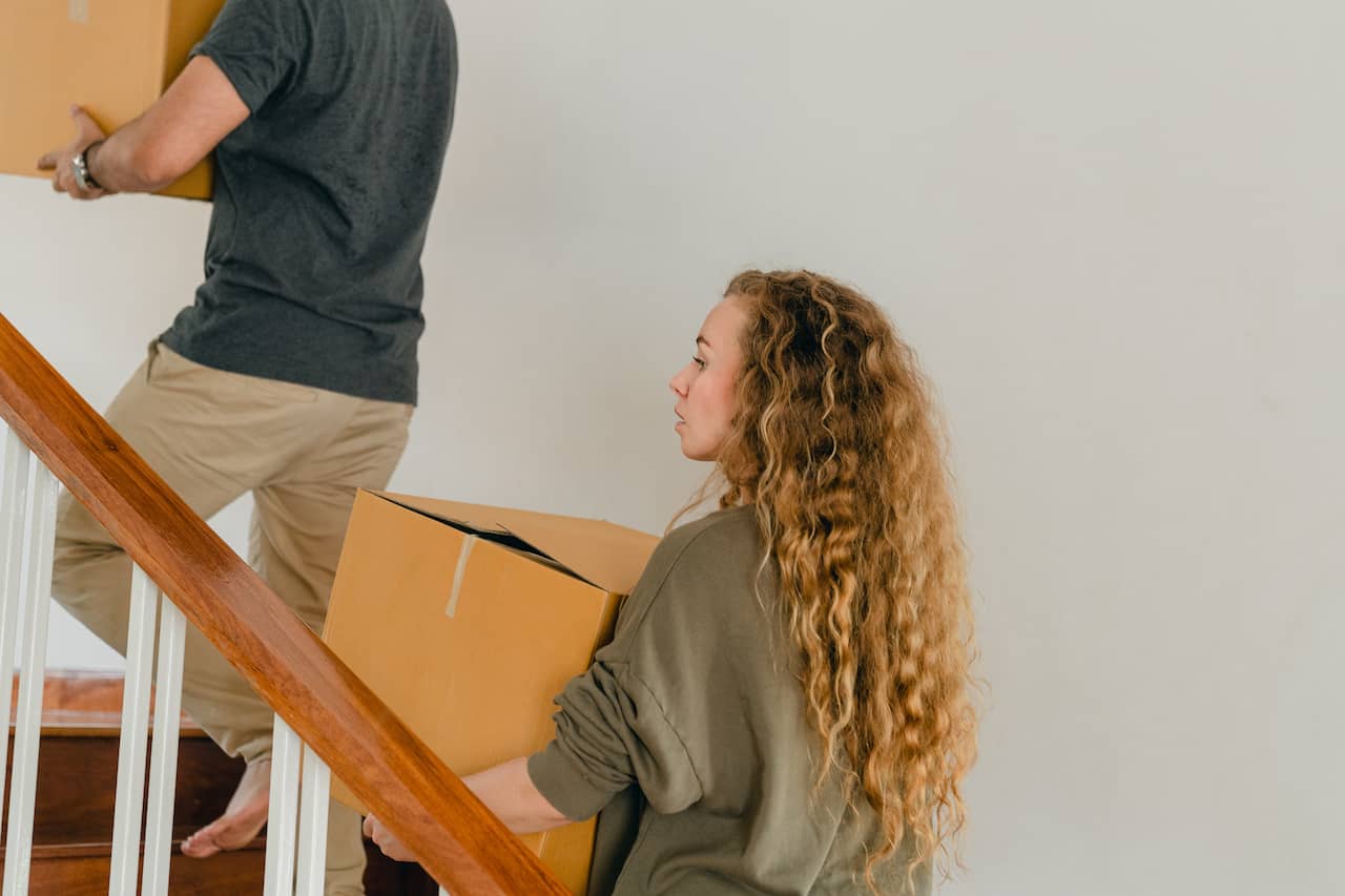 Two people carry cardboard moving boxes up an apartment staircase